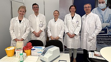 Mitarbeiter des Institutes für Klinische Chemie, inkl. Prof. Fischer, stehen in weißen Kitteln hinter einem Tisch, auf dem sich ein Cholesterin-Messgerät befindet.