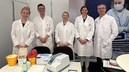 Mitarbeiter des Institutes für Klinische Chemie, inkl. Prof. Fischer, stehen in weißen Kitteln hinter einem Tisch, auf dem sich ein Cholesterin-Messgerät befindet.