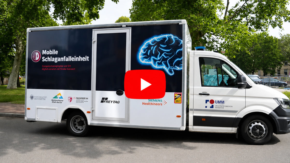 Mobile Schlaganfalleinheit der Universitätsmedizin Mannheim auf einer Straße unter Bäumen, mit leuchtendem Gehirnmotiv auf der Fahrzeugseite und rotem Play-Button in der Bildmitte.