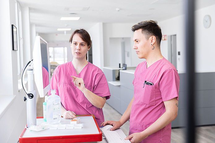 Pflegekraft und Pfleger im Klinikflur bedienen gemeinsam einen Computerwagen zur Dokumentation von Patientendaten.