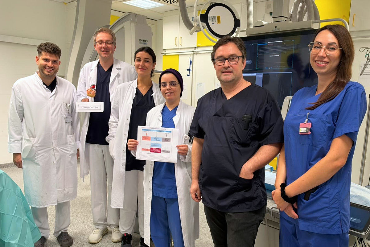 Team der Kardiologie der UMM in einem Herzkatheterlabor. Eine Mitarbeiterin hält eine Übersicht zur Studie, ein Kollege ein Modell mit der Aufschrift „Frauenherzen“.