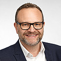 Ein Mann mit Brille lächelt vor hellem neutralem Hintergrund in die Kamera.