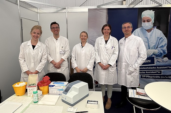 Fünf Personen in weißen Laborkitteln stehen hinter einem Tisch mit medizinischen Geräten und Verbrauchsmaterialien vor einem Messestand mit einem Banner.