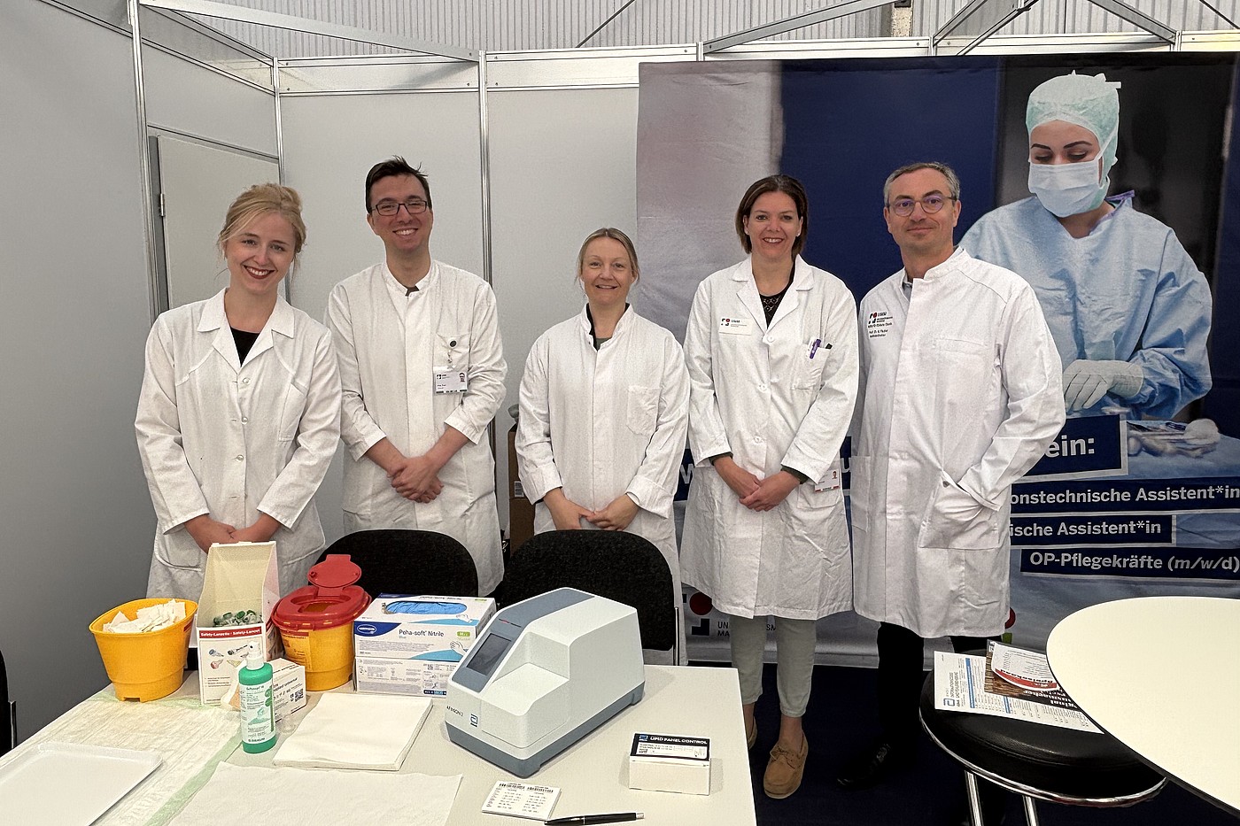 Fünf Personen in weißen Laborkitteln stehen hinter einem Tisch mit medizinischen Geräten und Verbrauchsmaterialien vor einem Messestand mit einem Banner.