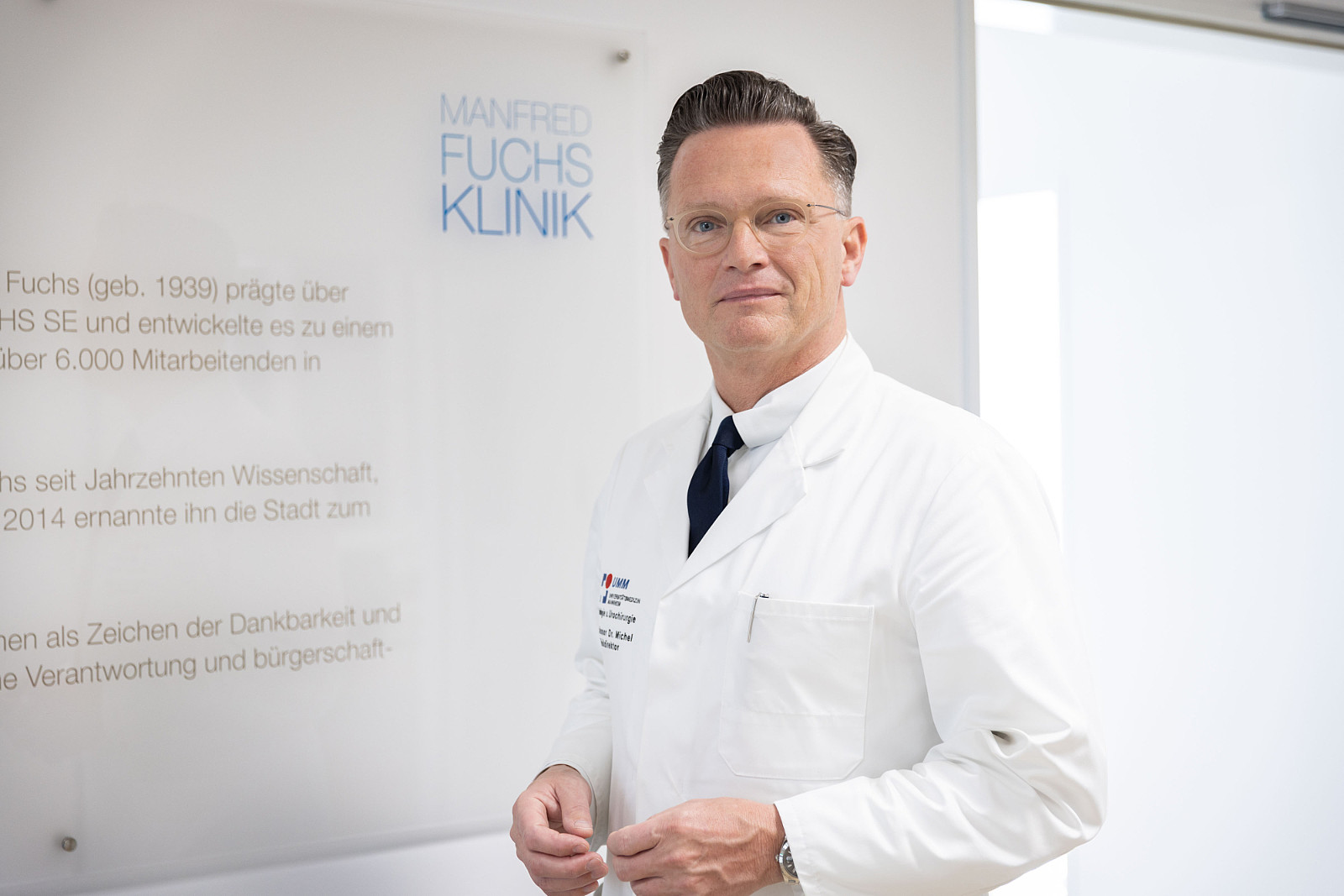 Professor Maurice Stephan Michel steht vor einer Wandtafel mit der Aufschrift „Manfred Fuchs Klinik“.