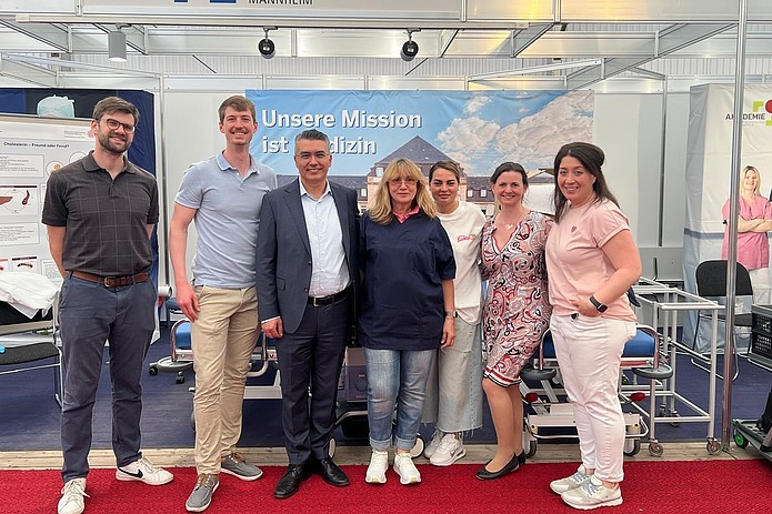 Sieben Personen stehen in einer Reihe vor einem Messestand mit dem Schriftzug 'UMM UNIVERSITÄTSMEDIZIN MANNHEIM' und einem Banner mit dem Text 'Unsere Mission ist Medizin'