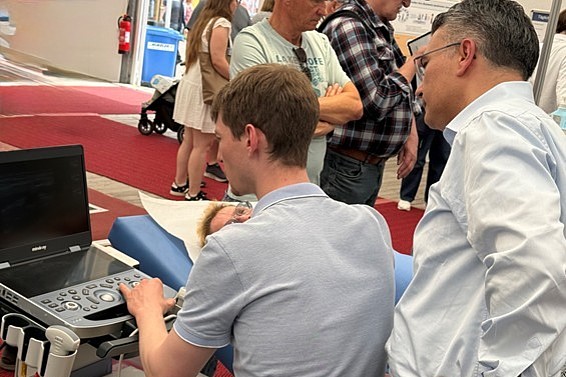 Junger Mann bedient medizinisches Ultraschallgerät vor mehreren stehenden Personen in einem Innenraum mit rotem Teppich