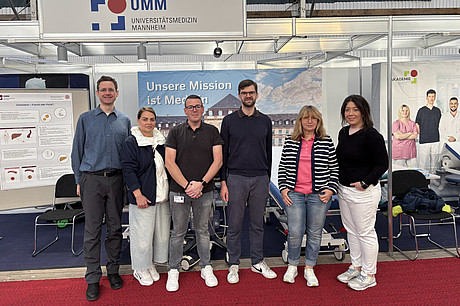 Sechs Personen stehen in einer Reihe vor einem Messestand mit dem Schild 'UMM UNIVERSITÄTSMEDIZIN MANNHEIM' und einem Banner mit dem Text 'Unsere Mission ist Mediz...'.