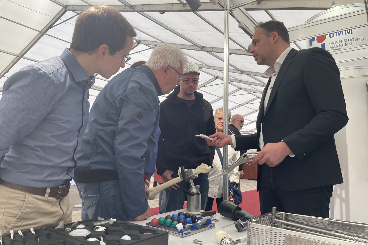 Mehrere Personen stehen an einem Tisch mit medizinischen Modellen und Instrumenten unter einem Zelt, eine Person im Anzug hält ein Modell eines Knochens mit einer Zange