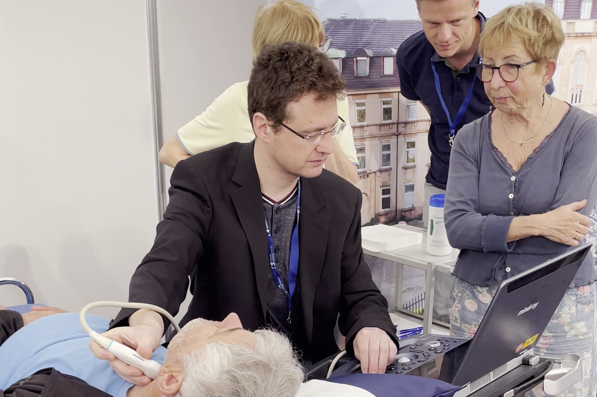 Person führt Ultraschalluntersuchung am Hals eines liegenden älteren Mannes durch, während zwei weitere Personen zuschauen