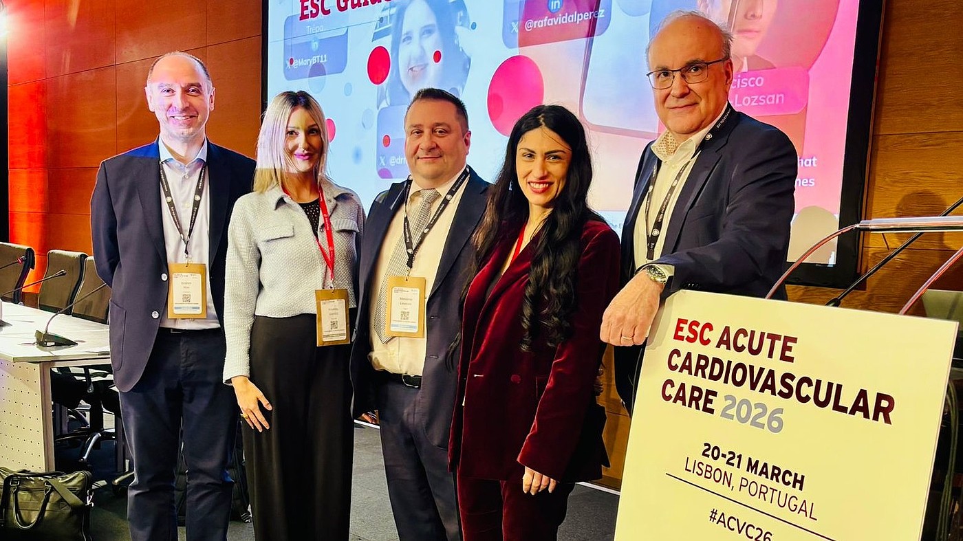 Fünf Personen mit Namensschildern stehen vor einer Bühne mit einem großen Bildschirm und einem Schild mit der Aufschrift 'ESC ACUTE CARDIOVASCULAR CARE 2026 20-21 MARCH LISBON, PORTUGAL'