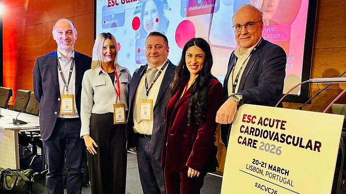 Fünf Personen mit Namensschildern stehen vor einer Bühne mit einem großen Bildschirm und einem Schild mit der Aufschrift 'ESC ACUTE CARDIOVASCULAR CARE 2026 20-21 MARCH LISBON, PORTUGAL'