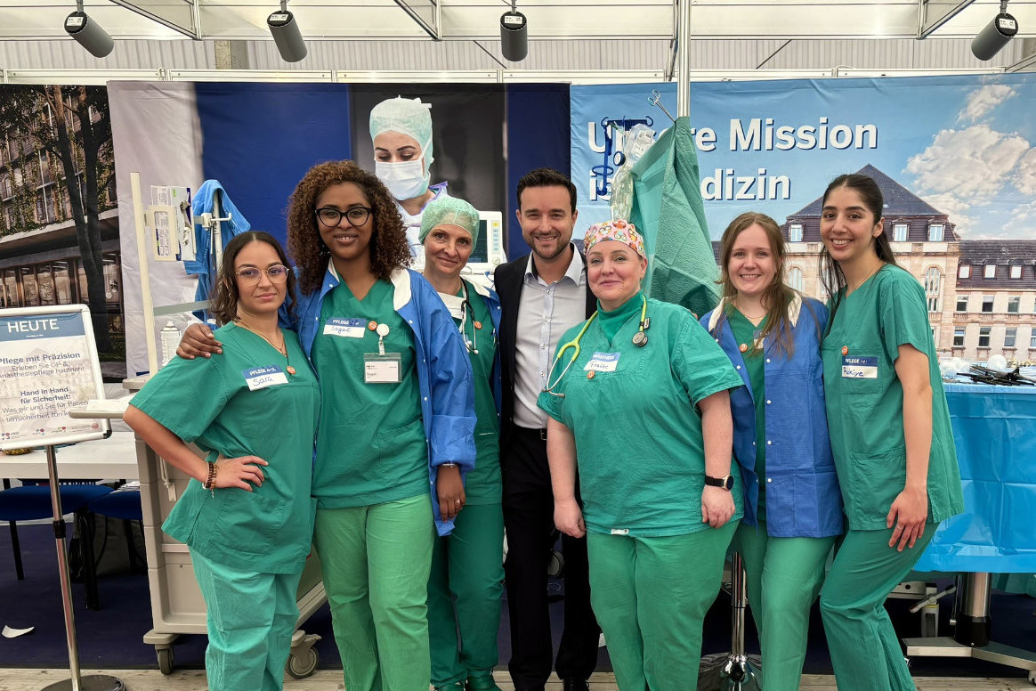 Gruppe von medizinischem Personal in grüner und blauer Arbeitskleidung vor einem Hintergrund mit Text und Gebäudefassade