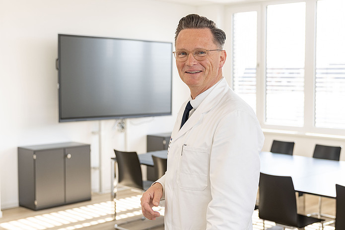 Professor Maurice Stephan Michel steht in einem Besprechungsraum vor einem Bildschirm und blickt in die Kamera.