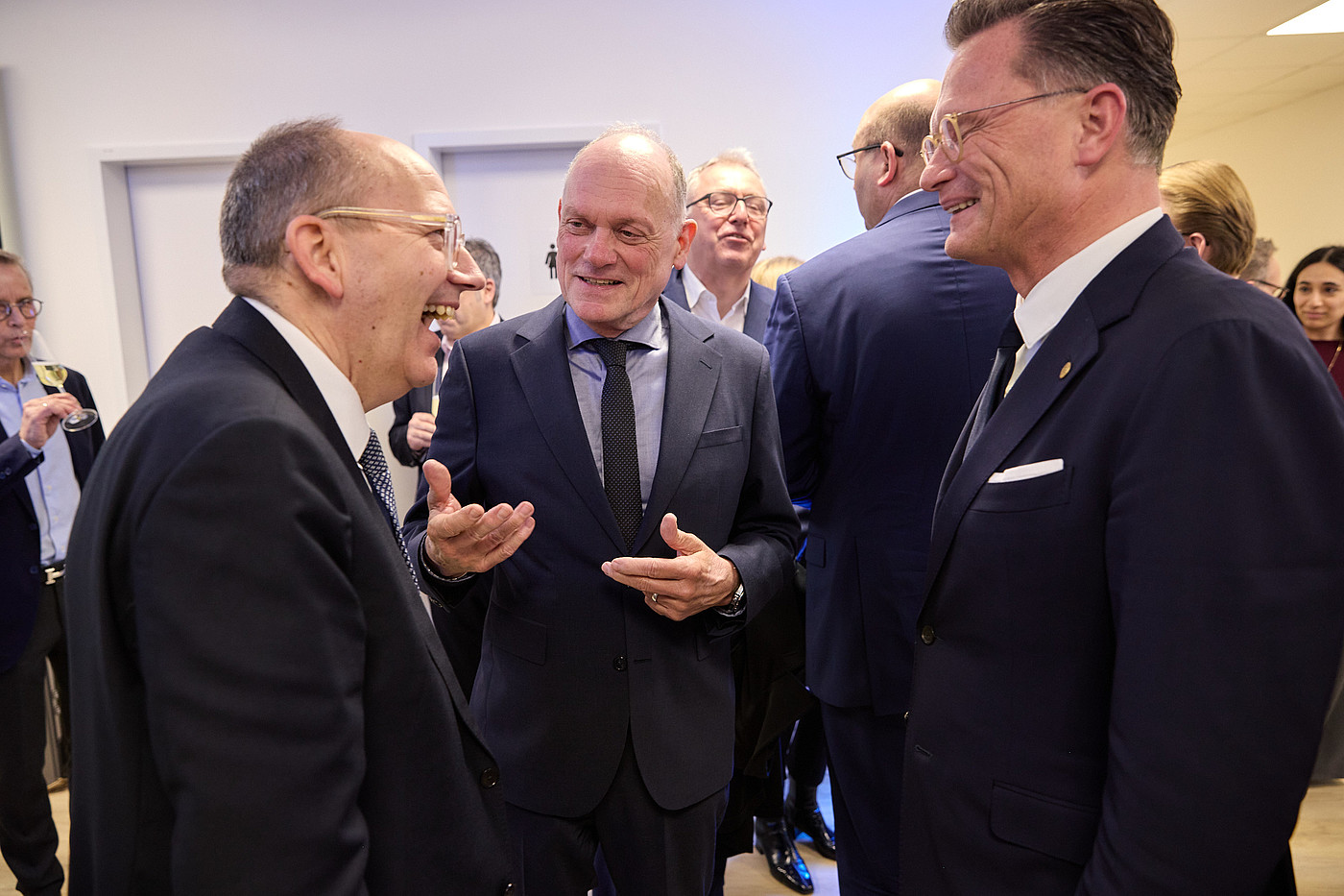 Drei Männer im Anzug stehen bei einer Veranstaltung zusammen und unterhalten sich in einem Foyer.