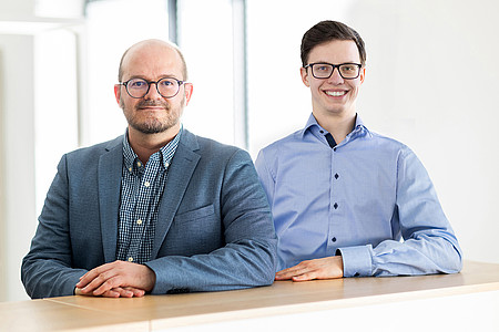 Das Foto zeigt zwei Männer, die nebeneinander hinter einer Mauer stehen, auf der sie jeweils ihren linken Unterarm abgelegt haben. Beide tragen Hemd ohne Krawatte, der linke Herr außerdem ein Jackett. Der linke Herr ist mittelalt und hat wenig Haar, der rechte Herr ist jung und hat braune Haare. Es handelt sich links um Professor Dr. Mayrhofer und rechts um Dominic Henning.
