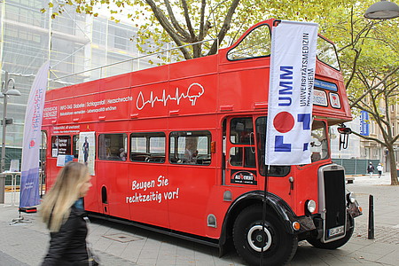 Ein roter Doppeldeckerbus mit der Aufschrift „Beugen Sie rechtzeitig vor!“ steht in einer Fußgängerzone vor dem Hintergrund eines Gebäudes mit Baugerüst. Der Bus wirbt im Rahmen der Initiative „Herzenssache Lebenszeit“ für Vorsorge bei Herz-Kreislauf-Erkrankungen, Diabetes und Schlaganfall. Daneben weht eine weiße Fahne mit dem Logo der Universitätsmedizin Mannheim. Im Vordergrund geht eine Frau vorbei.