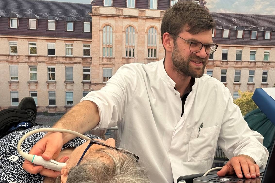 Mann in weißem Kittel führt Ultraschalluntersuchung am Hals einer liegenden älteren Frau durch, im Hintergrund ein historisches Gebäude