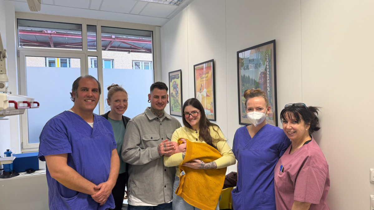 Sechs Personen in einer hellen Praxis: drei in blauer medizinischer Kleidung, drei Frauen. Eine der Frauen hält im Arm ein Säugling; alle lächeln in die Kamera, im Hintergrund Fenster und Bilder an der Wand.